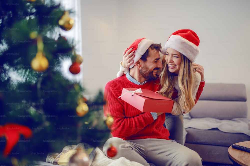 Overjoyed handsome bearded man sitting on sofa in living room, holding gift and hugging with his girlfriend. Both having santa hats on heads. In foreground is christmas tree. Living room interior.