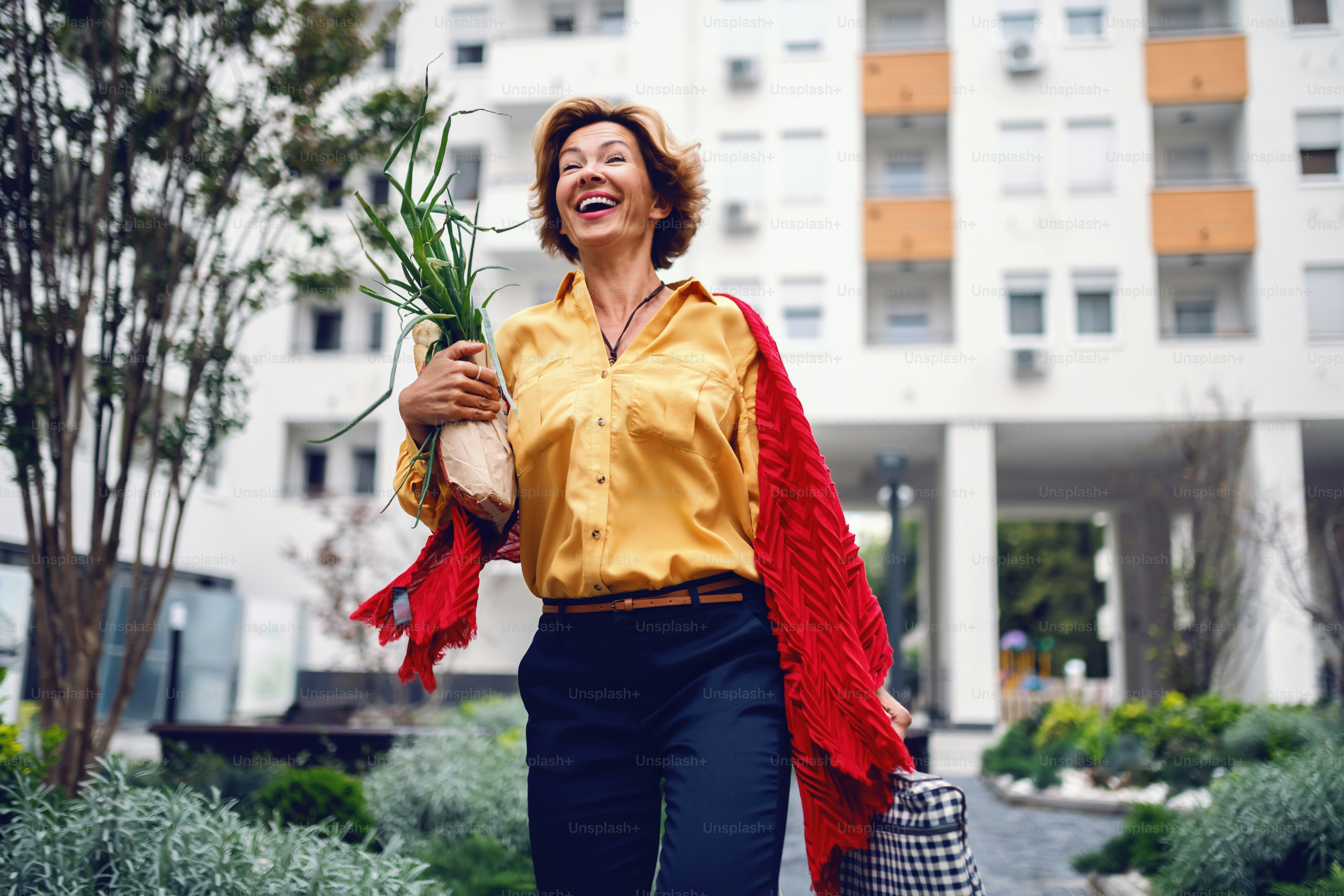Attractive caucasian blonde fashionable senior woman with scarf walking in park and carrying groceries. In background are buildings.