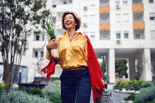 Attractive caucasian blonde fashionable senior woman with scarf walking in park and carrying groceries. In background are buildings.