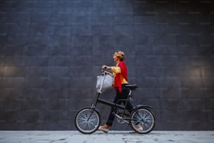 ゴージャスで健康的な金髪の白人シニア女性の全長は、自転車を押して灰色の壁を通り過ぎます。