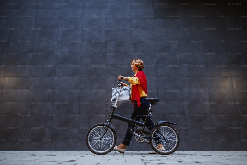 Full length of gorgeous healthy blonde caucasian senior woman pushing bicycle and passing by gray wall.