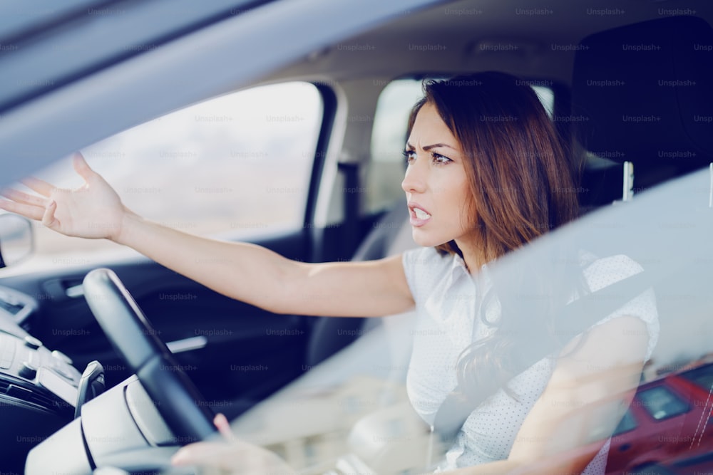 Angry attractive caucasian brunette yelling at other drivers while sitting in car.