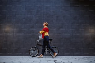 Full length of gorgeous healthy blonde caucasian senior woman pushing bicycle and passing by gray wall.