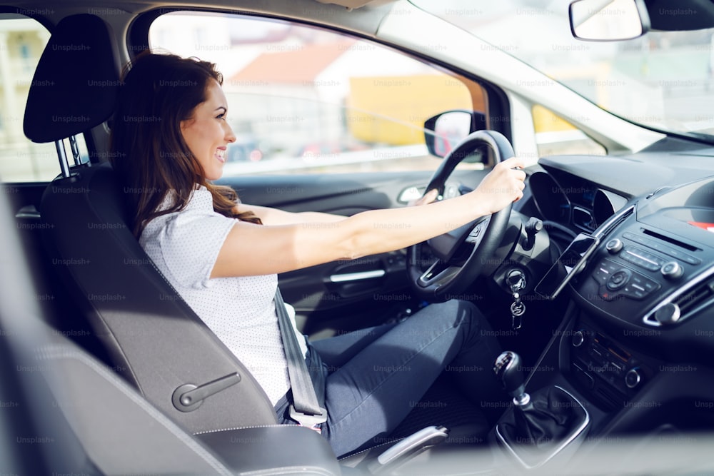 Side view of cheerful caucasian brunette in shirt driving her car. Hands are on steering wheel and safety belt is buckled on.