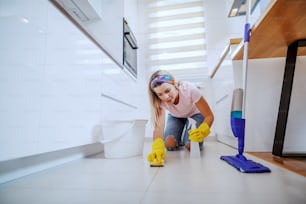 Ordentliche kaukasische blonde Hausfrau mit Gummihandschuhen, die in der Küche kniet und den Küchenboden mit Schwamm und Sprühgerät putzt.
