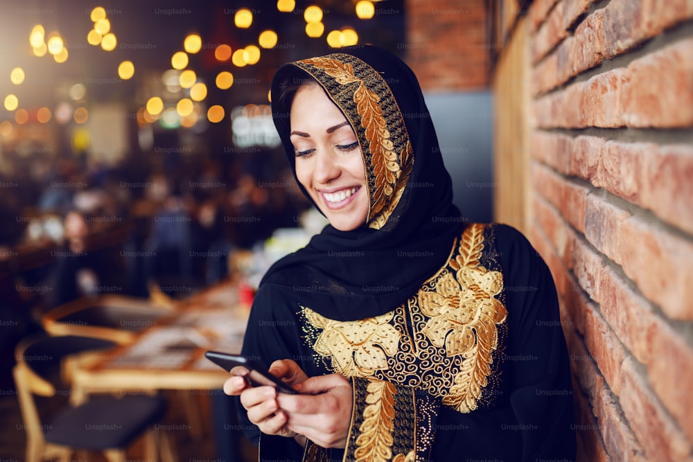 Portrait of beautiful smiling muslim woman dressed in traditional wear sitting in coffee shop and using smart phone for sending or reading message.