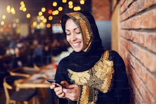 Ritratto di bella donna musulmana sorridente vestita in abiti tradizionali seduti in caffetteria e utilizzando lo smartphone per l'invio o la lettura del messaggio.