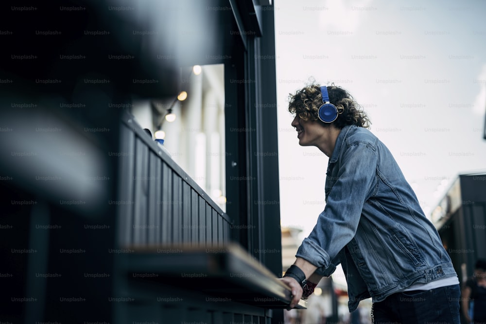 Merry curly man in denim clothes is standing in earphones at kiosk window. Website banner