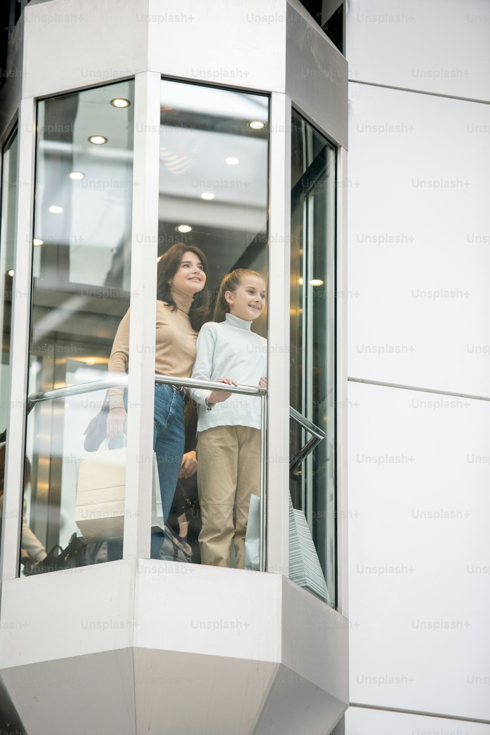 Pretty young woman and girl in casualwear looking through elevator window while moving upwards during shopping