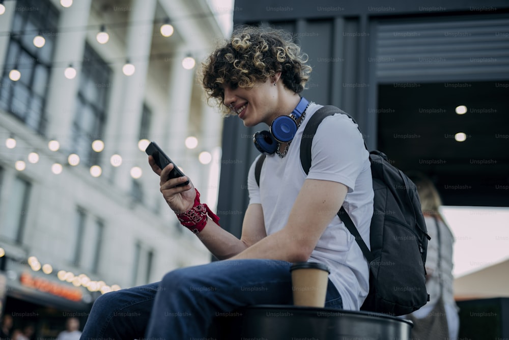 Merry curly young man with headphones is sitting in city and using mobile phone