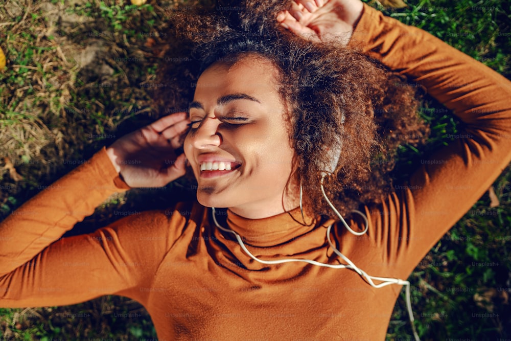 Beautiful mixed race young woman with curly hair and in turtleneck lying down on lawn in park and enjoying music.