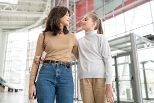 Menina bonita e sua mãe abraçada olhando uma para a outra enquanto se movem pelo grande shopping e conversam