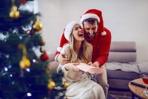 Stupita donna bionda caucasica sveglia seduta sul divano in soggiorno e ricevendo un regalo dal suo ragazzo. Entrambi hanno cappelli da Babbo Natale in testa. In primo piano c'è l'albero di Natale. Interno del soggiorno.