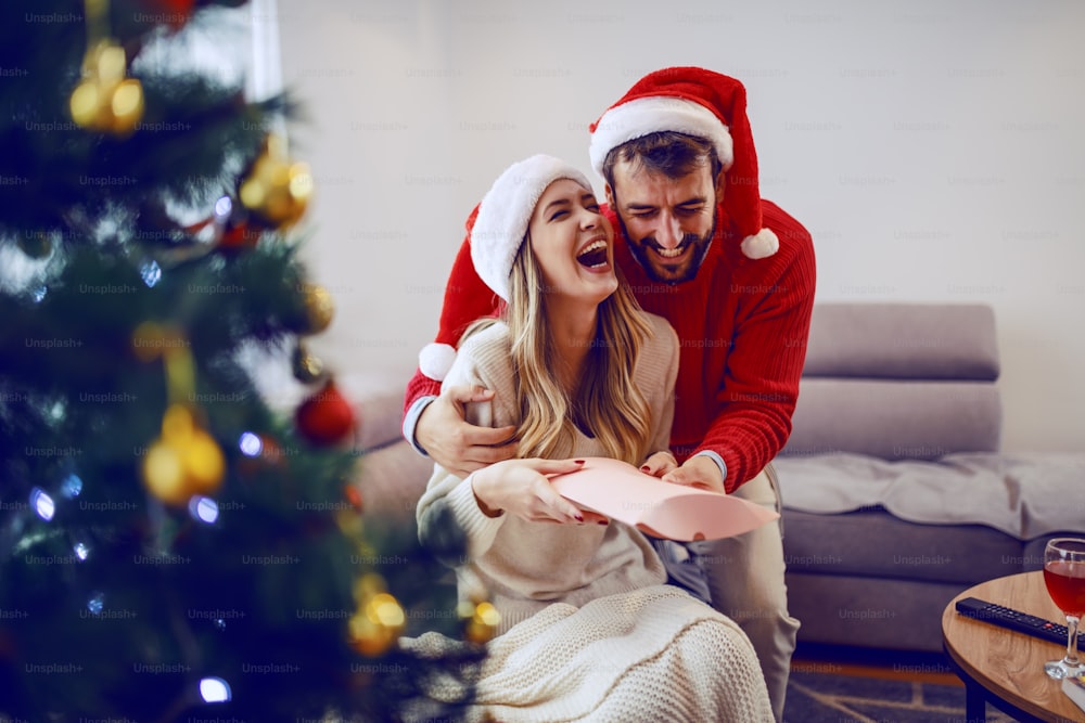 Amazed cute Caucasian blonde woman sitting on sofa in living room and receiving gift from her boyfriend. Both having santa hats on heads. In foreground is christmas tree. Living room interior.