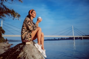 Vue latérale de charmante brune en robe fleurie et avec bandeau assis sur un rocher sur le rivage et buvant une boisson dans une tasse. En arrière-plan se trouve le pont.