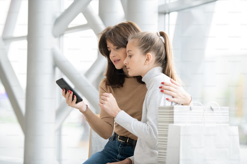 Young mother showing something curious to her daughter in smartphone while discussing it with her after shopping