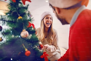 Happy caucasian young couple with santa hats on heads decorating christmas tree while standing in living room.