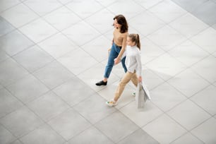 캐주얼한 젊은 여성과 그녀의 딸은 쇼핑을 하는 동안 현대적인 쇼핑몰을 따라 움직이는 종이 가방을 들고 있다