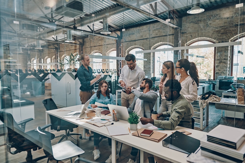 Group of young modern people in smart casual wear communicating and using modern technologies while working in the office