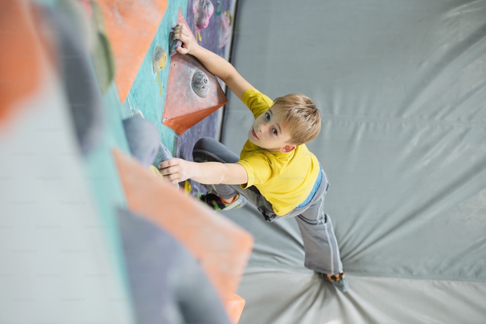 Concentrated schoolboy looking at you while standing on grey mat and keeping one foot on rock of climbing equipment during training