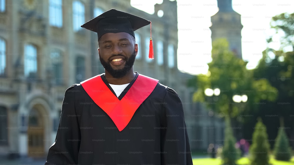 Studente afro-americano felice in abito di laurea sinceramente sorridente alla telecamera
