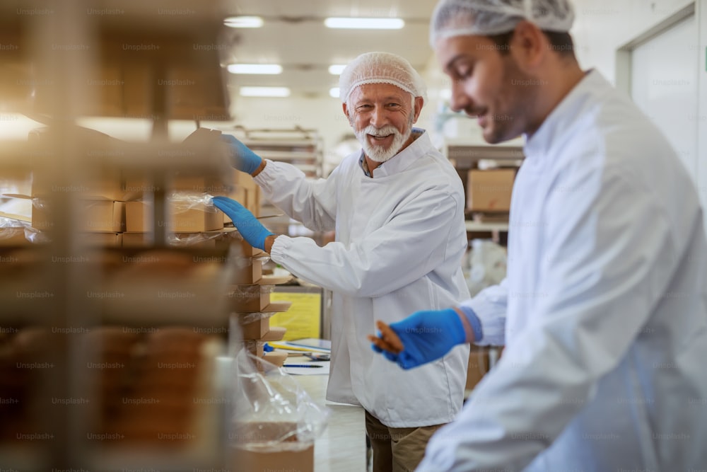Cheerful Caucasian employees dressed in white sterile uniforms packing cookies in boxes while standing in food plant.