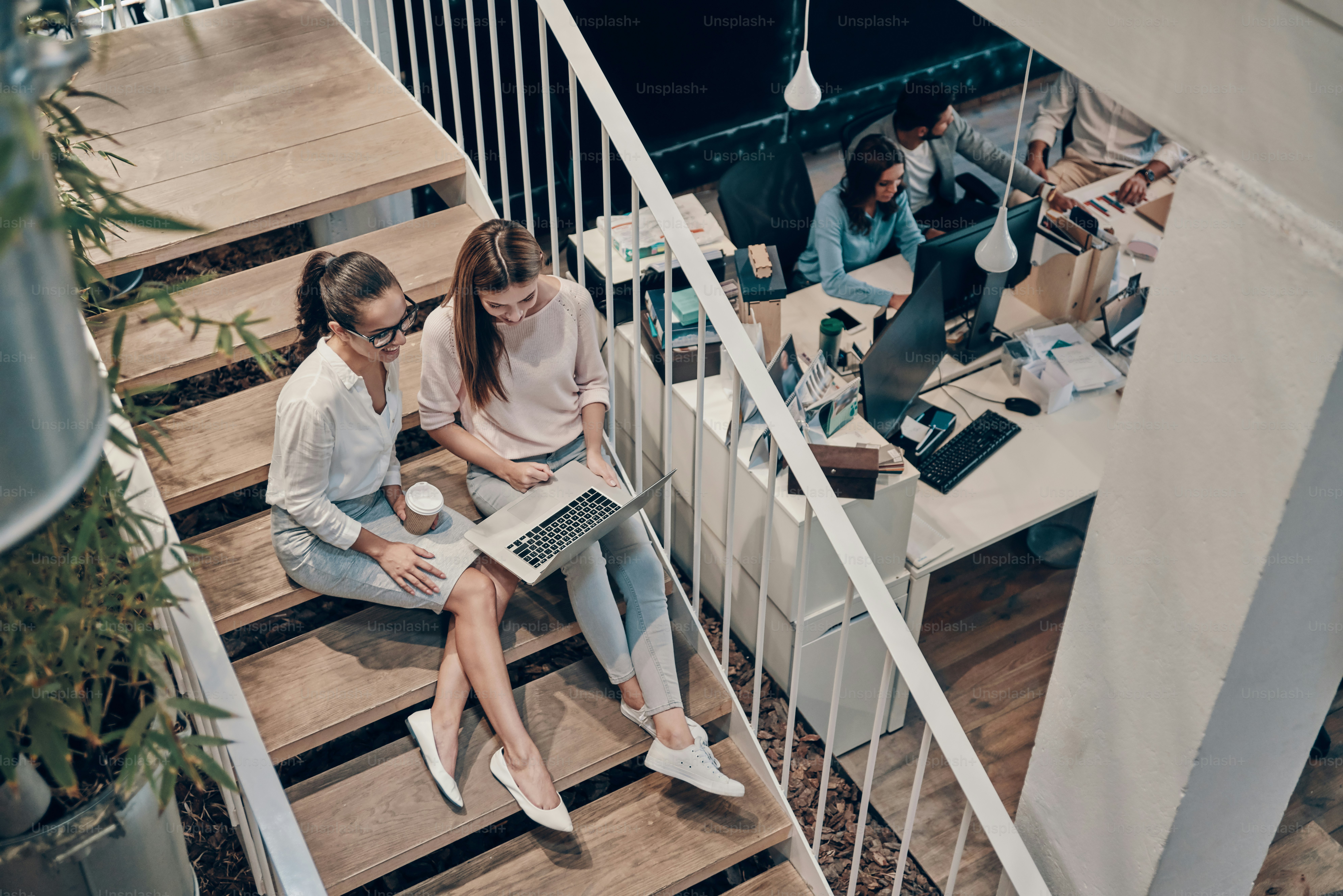 Top view of two young modern women in smart casual wear communicating and using laptop while sitting on the stairs