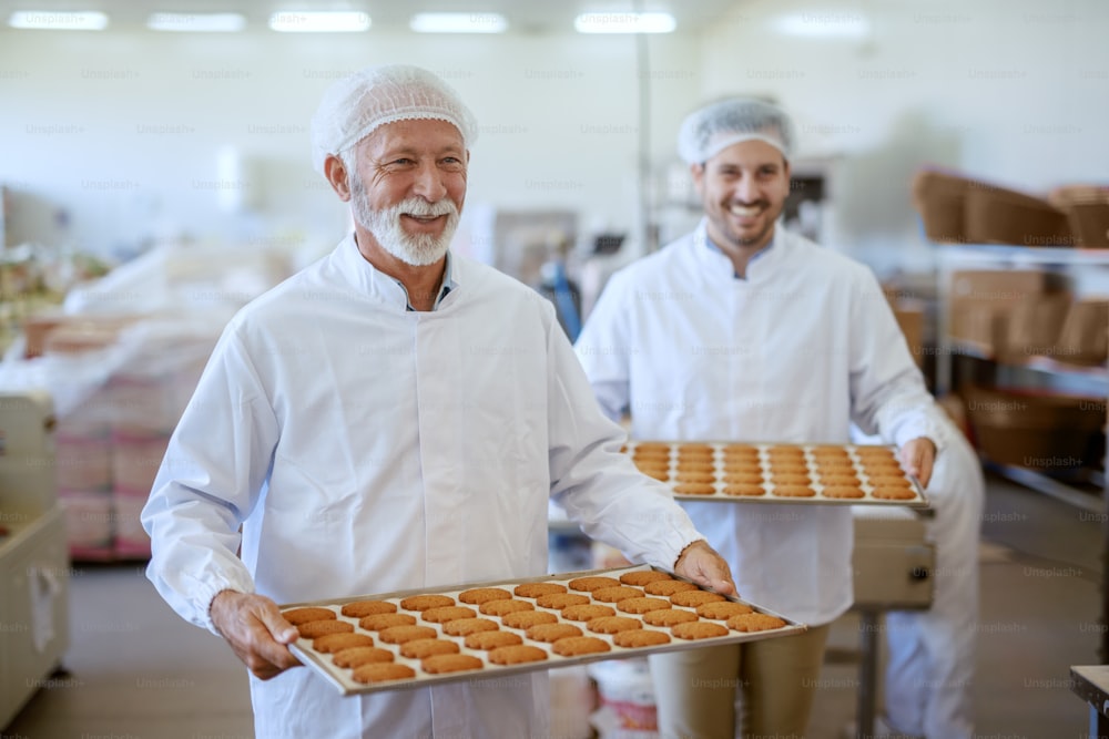Dos alegres empleados caucásicos que llevan bandejas con galletas frescas. Ambos están vestidos con uniformes blancos estériles y con redes para el cabello. Interior de la planta de alimentos.