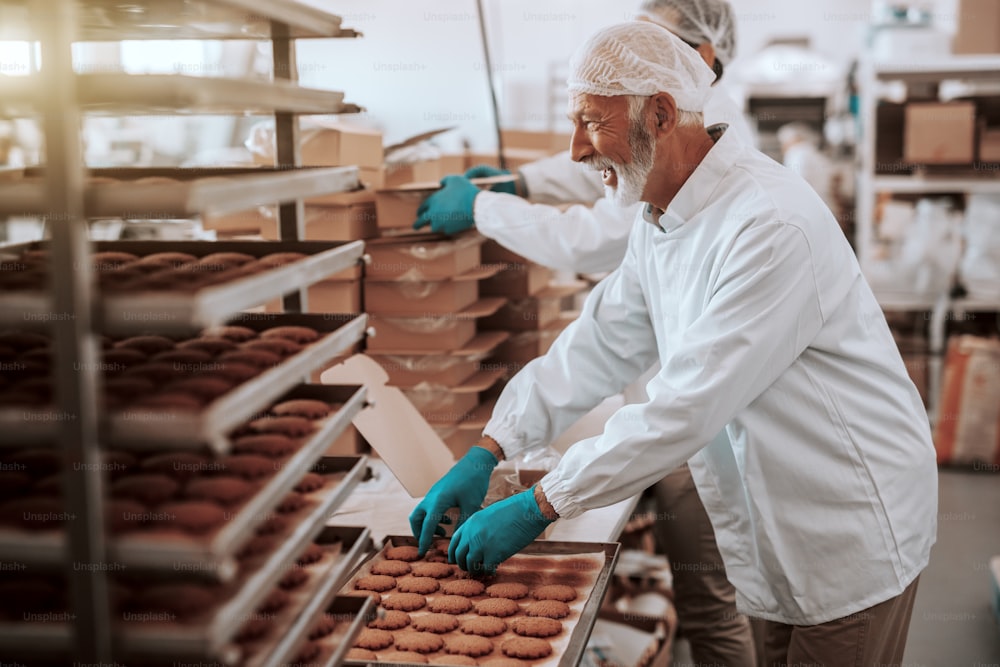 Dois funcionários caucasianos dedicados e esforçados, vestidos com uniformes brancos estéreis, coletando e embalando biscoitos em caixas. Interior da planta alimentícia.