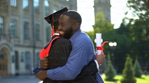 Pai afro-americano extremamente orgulhoso abraçando filho formatura com diploma, alegria