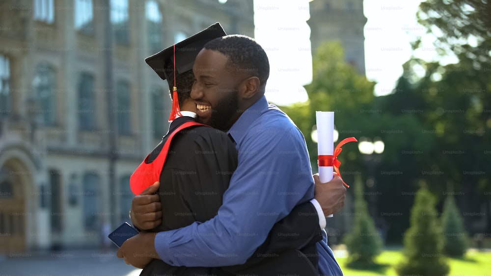 Padre afroamericano extremadamente orgulloso abraza a su hijo graduado con diploma, alegría