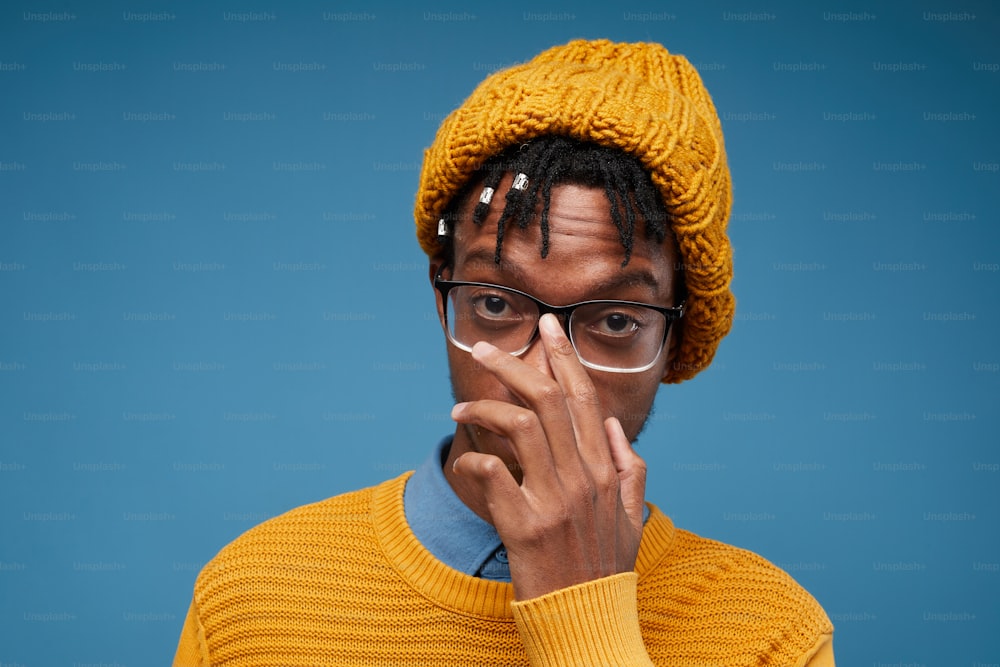Portrait of trendy African-American man wearing bright knit hat and sweater posing against blue background adjusting hid glasses, copy space