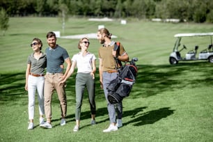 Des amis jeunes et élégants marchant avec du matériel de golf, traînant ensemble sur le magnifique parcours avec une voiture de golf en arrière-plan