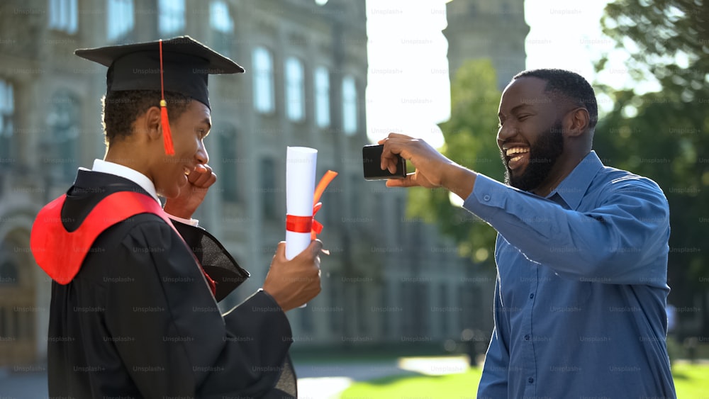 卒業証書を持つ幸せな卒業息子のスマートフォンビデオを作る笑顔のお父さん、イベント
