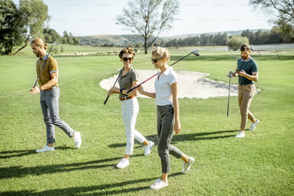 Junge Freunde, die lässig gekleidet sind, gehen während eines Spiels an einem sonnigen Tag mit Puttern auf dem Golfplatz spazieren