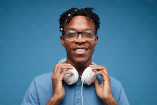 Head and shoulders portrait of contemporary African-American man wearing headphones smiling at camera while posing against blue background, copy space
