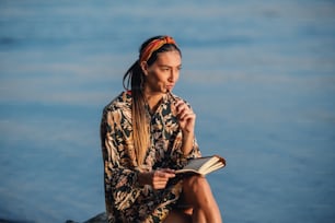 Portrait of pensive attractive caucasian brunette in floral dress and with headband sitting on rock on shore next to river and writing diary.