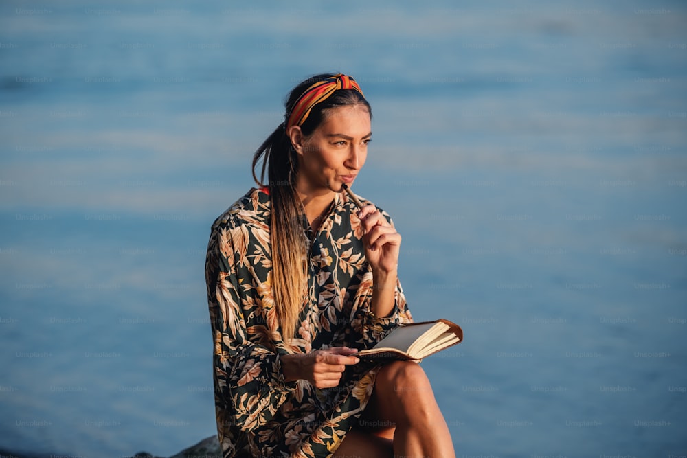 Portrait d’une jolie brune caucasienne pensive en robe à fleurs et avec bandeau assis sur un rocher sur le rivage à côté de la rivière et écrivant un journal intime.