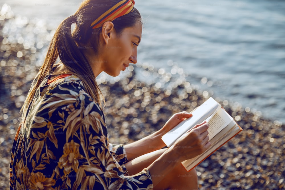 Morena caucasiana atraente em vestido sentado perto do rio em dia ensolarado, relaxando e lendo livro.