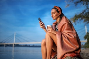 Attractive caucasian brunette covered with blanket sitting on the shore on rock, holding mug and using smart phone for reading or writing message.