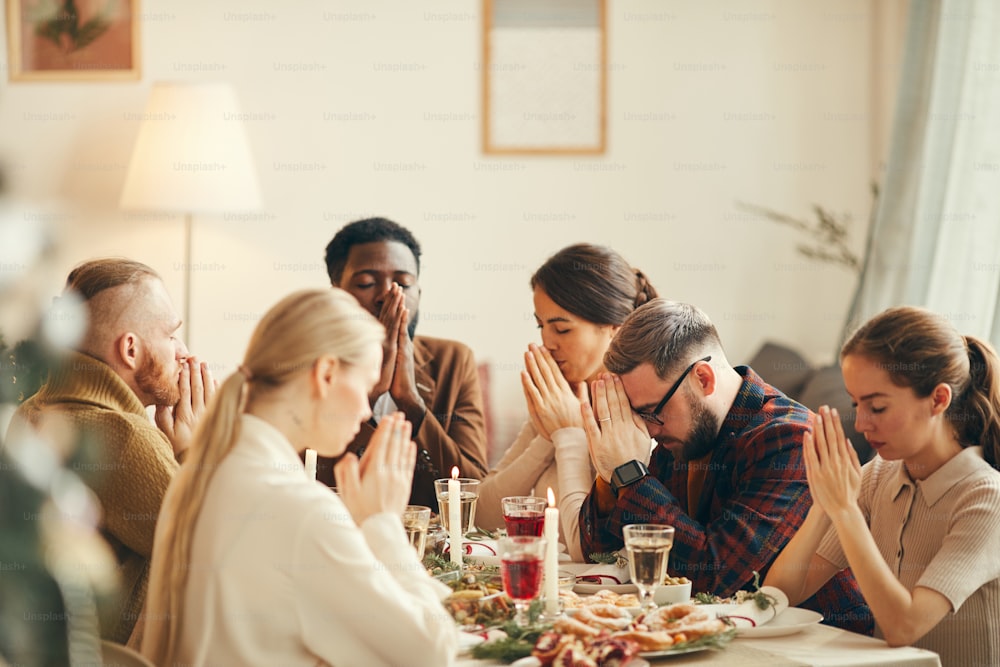 Gruppo multietnico di persone che dicono grazia a tavola durante il banchetto di Natale con amici e familiari