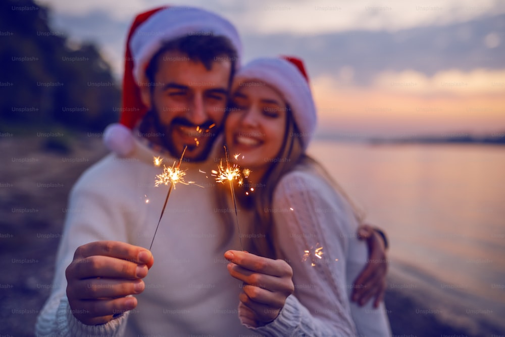 Overjoyed caucasian couple in love standing on coast near river and holding sparklers. Both are dressed in white sweaters and having santa hats on heads. Selective focus on sparklers.