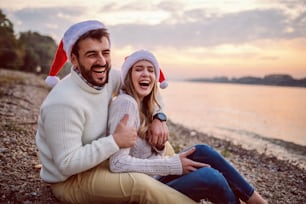 Happily in love Caucasian couple sitting on coast next to river. Man hugging woman. Both are dressed in white sweaters and having santa hats in heads.