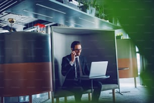 Smiling handsome caucasian businessman in suit and with eyeglasses sitting at workplace, having business call and looking at laptop.