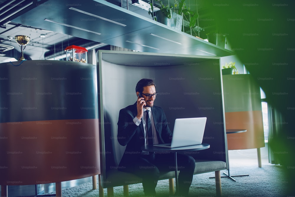 Smiling handsome caucasian businessman in suit and with eyeglasses sitting at workplace, having business call and looking at laptop.