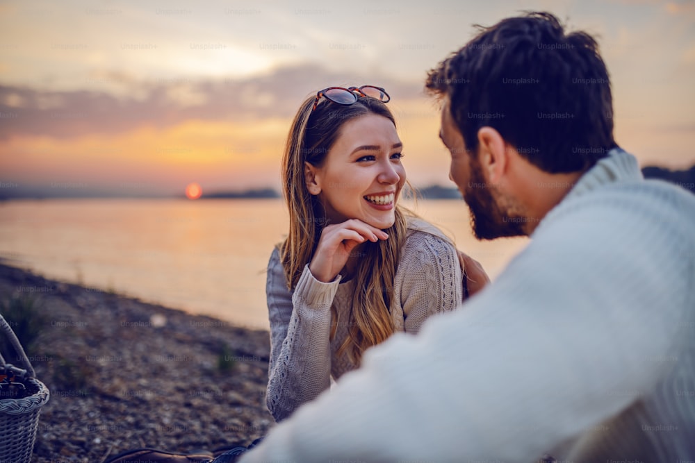 Linda morena caucásica sonriente sentada en la costa cerca del río con su amado novio y coqueteando. En el fondo está la puesta de sol.