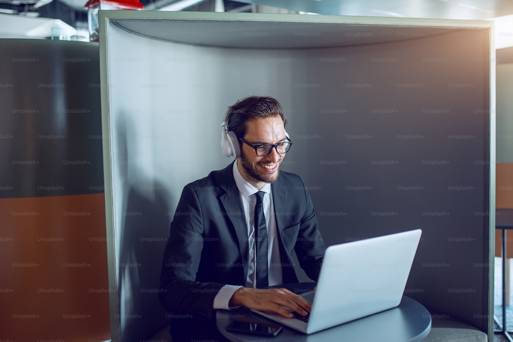 Sorridente bell'uomo d'affari barbuto caucasico in giacca e cravatta, con occhiali e cuffie seduto sul posto di lavoro e digitando sul laptop.