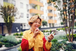 Retrato de una hermosa mujer mayor rubia caucásica sosteniendo el espejo y poniéndose lápiz labial mientras está parada en el parque.