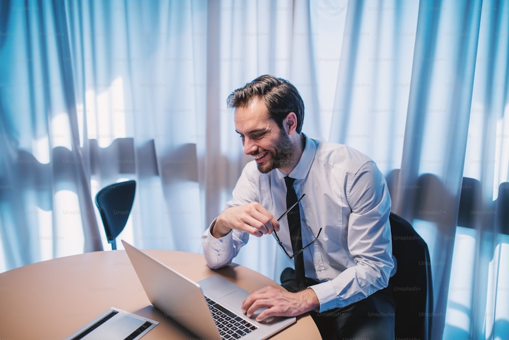 Ambitious caucasian elegant bearded businessman in shirt and tie holding eyeglasses and using laptop while sitting in office.