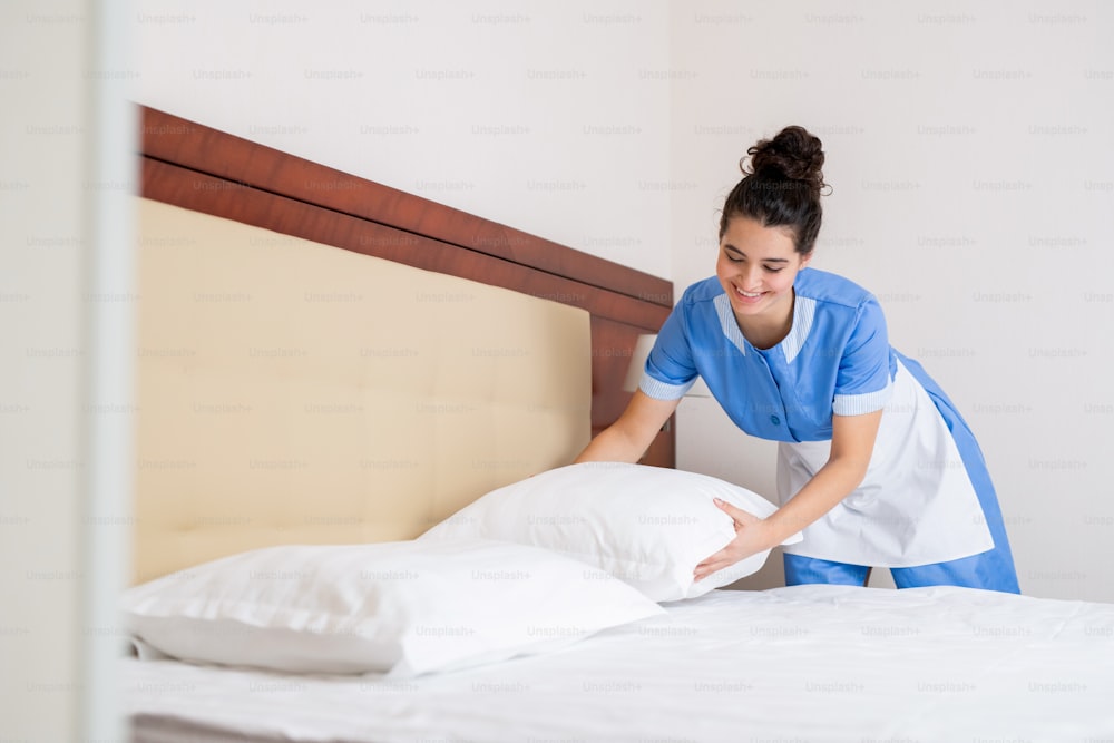 Happy brunette girl in uniform of chamber maid putting white clean pillows on bed while doing her work in hotel room